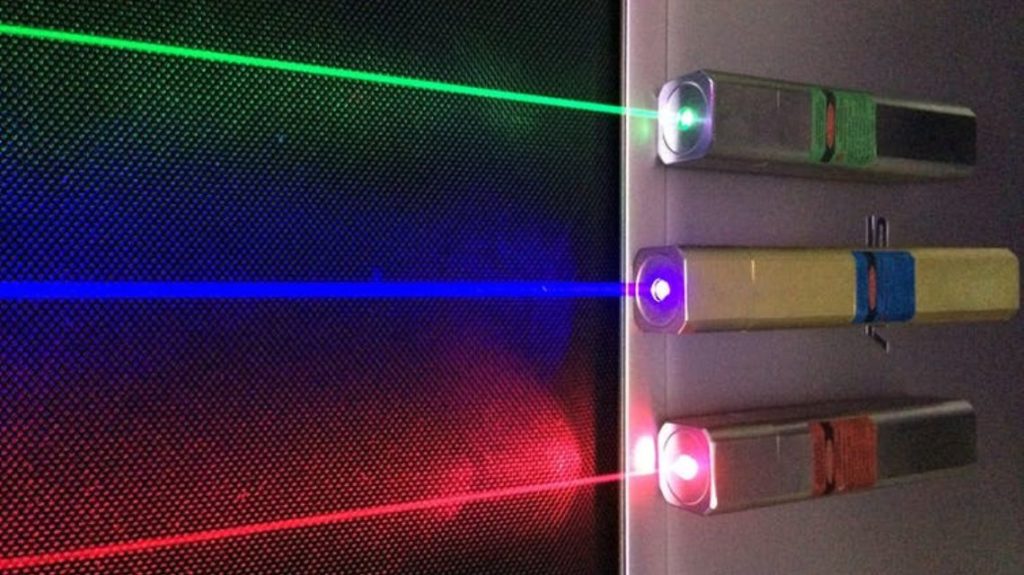 Top right corner: Grey laser pointer with a green strip around the middle, beaming a green light in front of a black screen.

Below the grey laser: Golden laser pointer with a blue strip around the middle, beaming a blue light in front of a black screen.

Below the golden laser: Grey laser pointer with a red strip around the middle, beaming a red light in front of a black screen.
