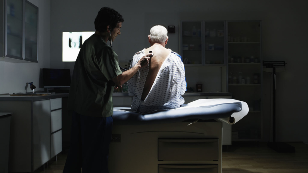 Older men sitting in hospital bed while a physician uses a stetoscope on his back